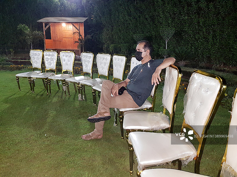 一名男子戴着口罩，晚上独自坐在草坪的椅子上。图片素材