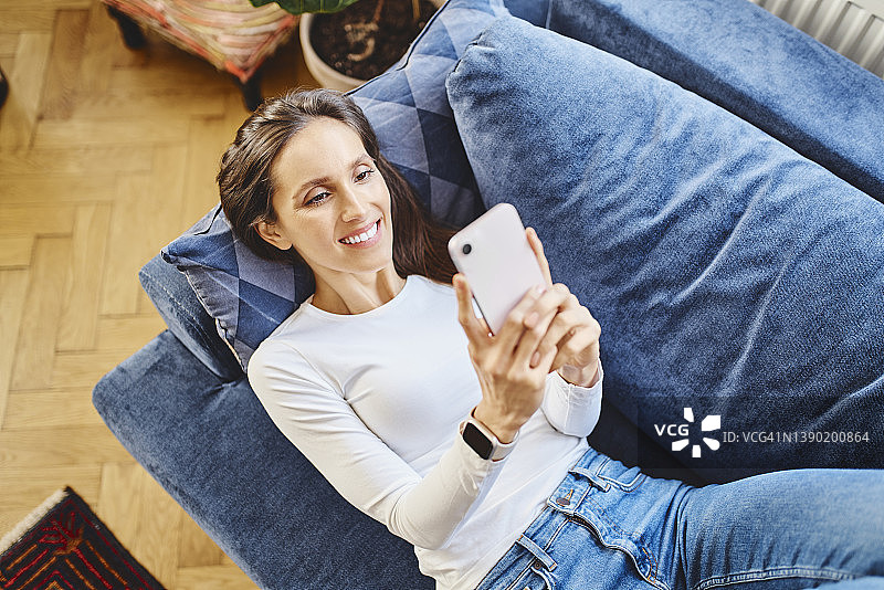 快乐的女人躺在客厅的沙发上通过智能手机上网图片素材