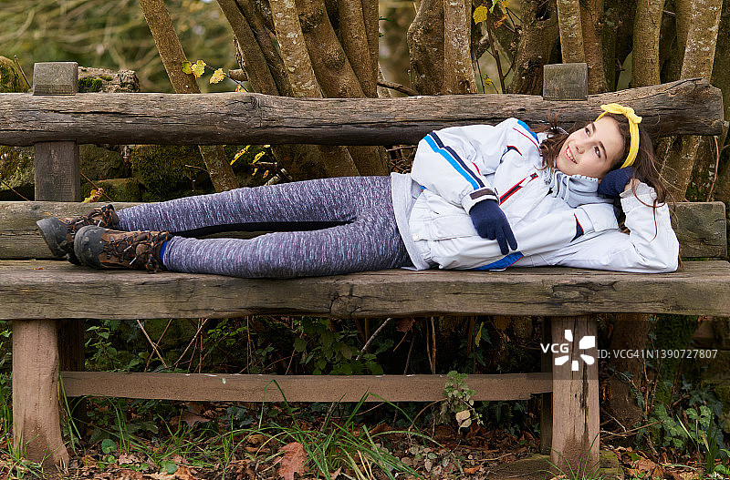 美丽的女孩穿着暖和的衣服，躺在用回收的木头做的质朴的长凳上，看起来沉思和快乐。图片素材