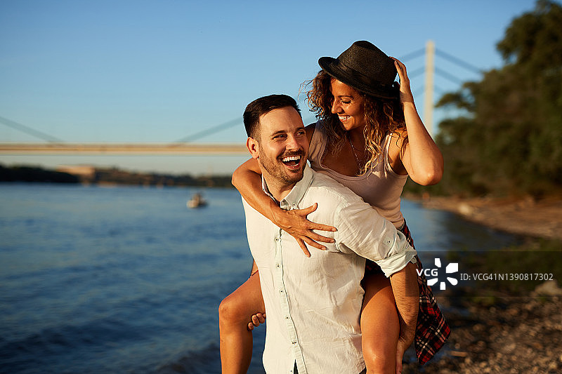 一个微笑的年轻人在河边背他正在笑的女朋友图片素材