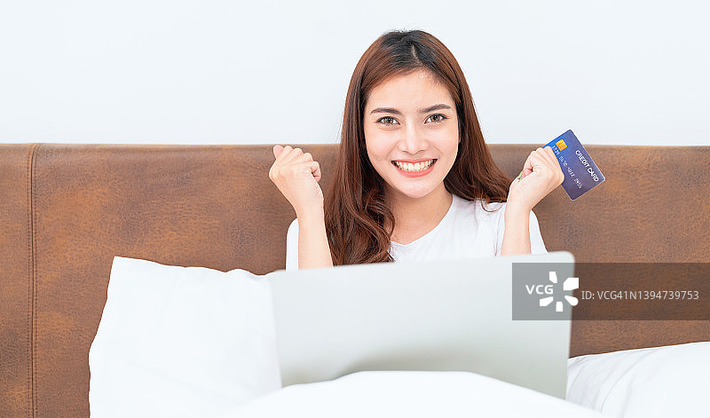 快乐的亚洲女人在家里网购，女人在家里的床上用信用卡和笔记本电脑网购图片素材