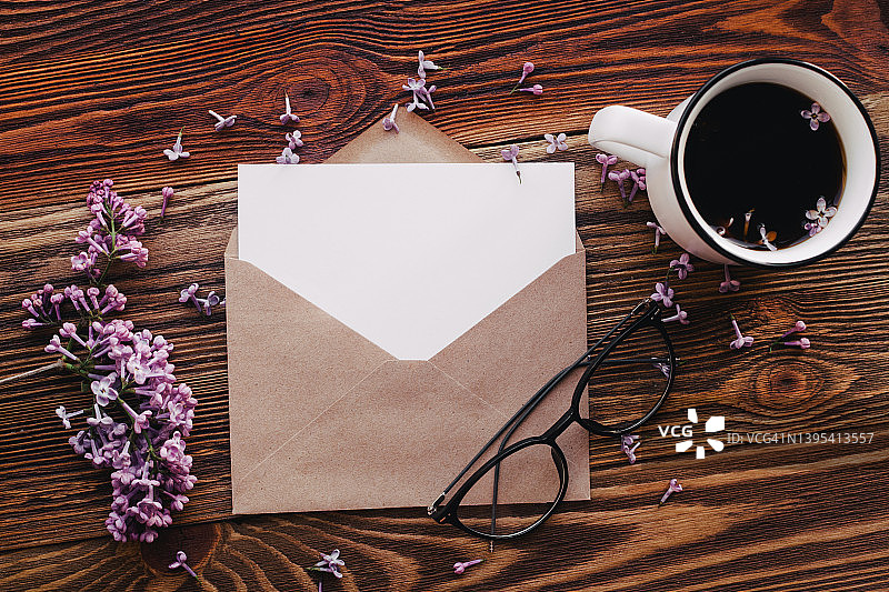 紫丁香紫色的花在棕色的木头背景，信封和空白纸卡的拷贝空间。一杯咖啡，一副眼镜。问候春夏贺卡。粉色花母亲节卡片。图片素材