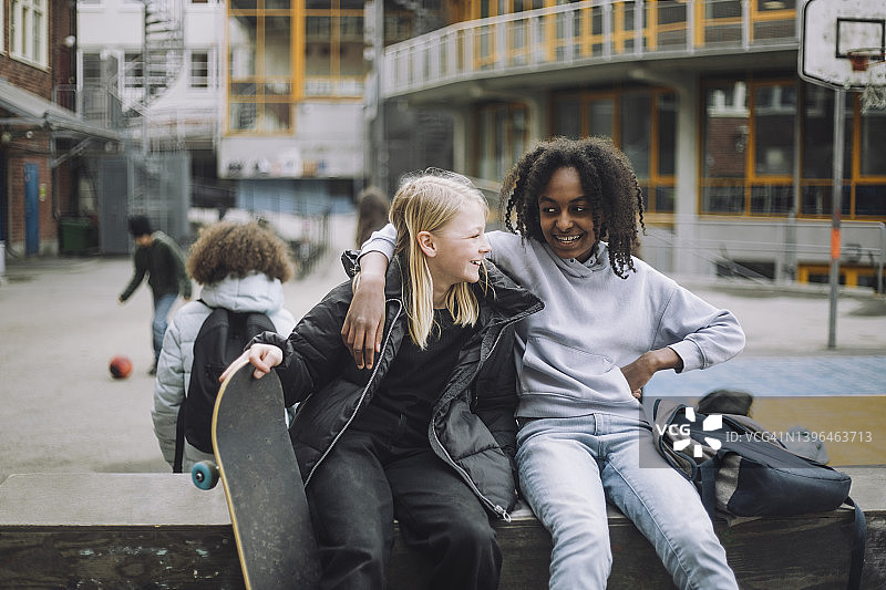 微笑的女孩拿着滑板，而坐在与朋友在挡土墙在学校校园图片素材