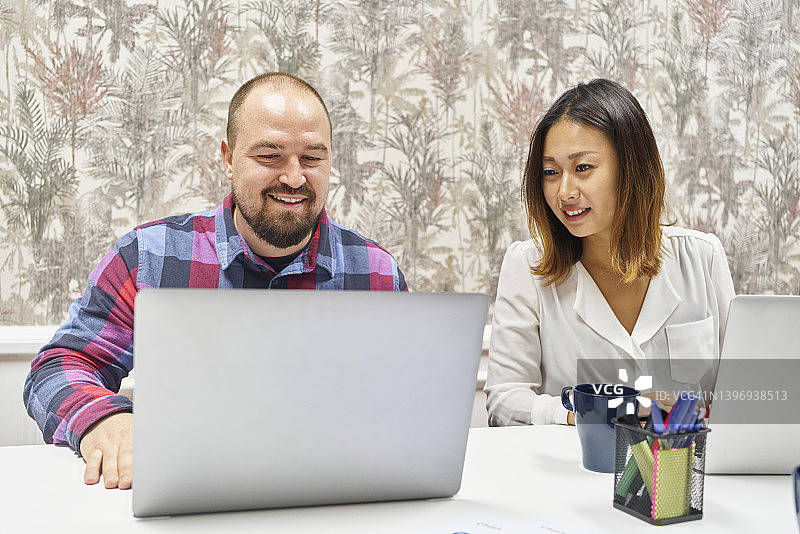 白人男性坐在旁边的亚洲女性同事在办公室会议桌上使用笔记本电脑的前视图图片素材