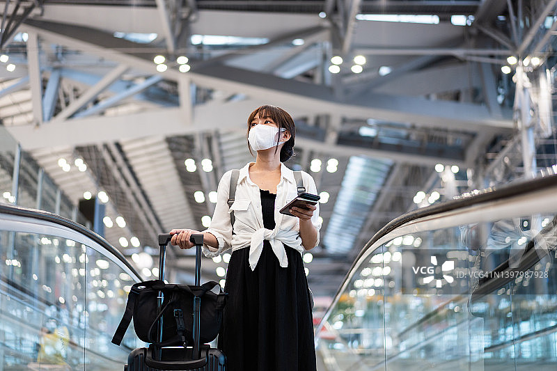 一名戴口罩的亚洲年轻女子站在机场候机楼的自动扶梯上打电话图片素材