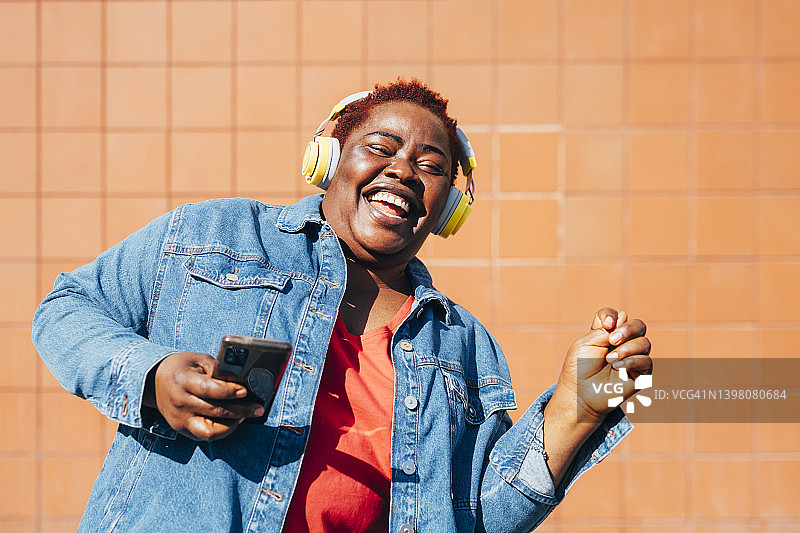 快乐的女人拿着智能手机，通过无线耳机听音乐，在墙壁前享受图片素材