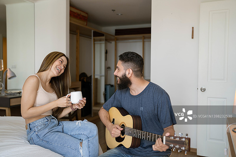 幸福的年轻的多民族夫妇喜欢唱歌和吉他，而坐在卧室在早上。图片素材