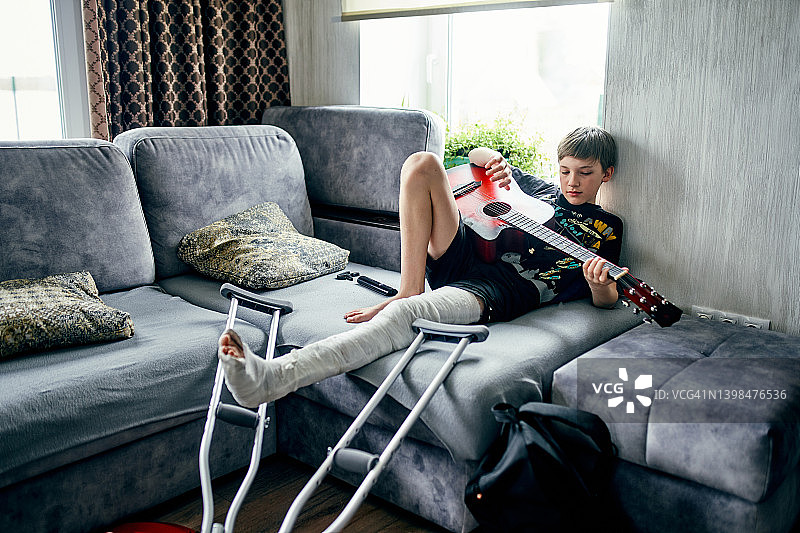 一个12岁的男孩断了腿，打了石膏，坐在家里的沙发上弹着红色的吉他。图片素材
