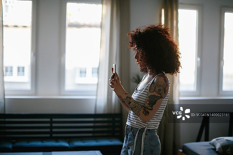 年轻的卷发纹身女子在家里用智能手机图片素材