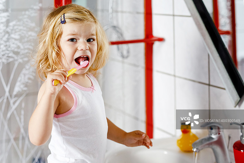 可爱可爱的学步小女孩抱着牙刷在浴室里刷第一颗牙。漂亮的宝宝正在学习清洁乳牙。儿童晨间健康卫生常规图片素材