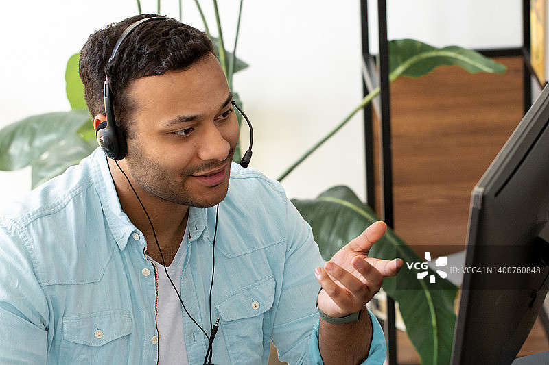 印度男性呼叫中心代理戴着耳机工作在支持热线，同时坐在现代办公室肖像亚洲帮助台工作人员通过耳机使用视频通话与客户交谈图片素材