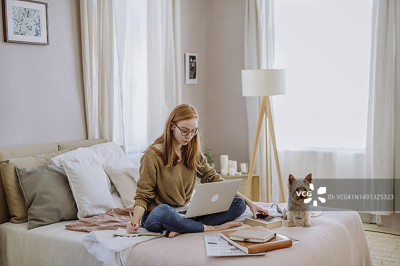 在家里的床上，宠物狗旁边，坐在笔记本电脑旁的女人在写书图片素材