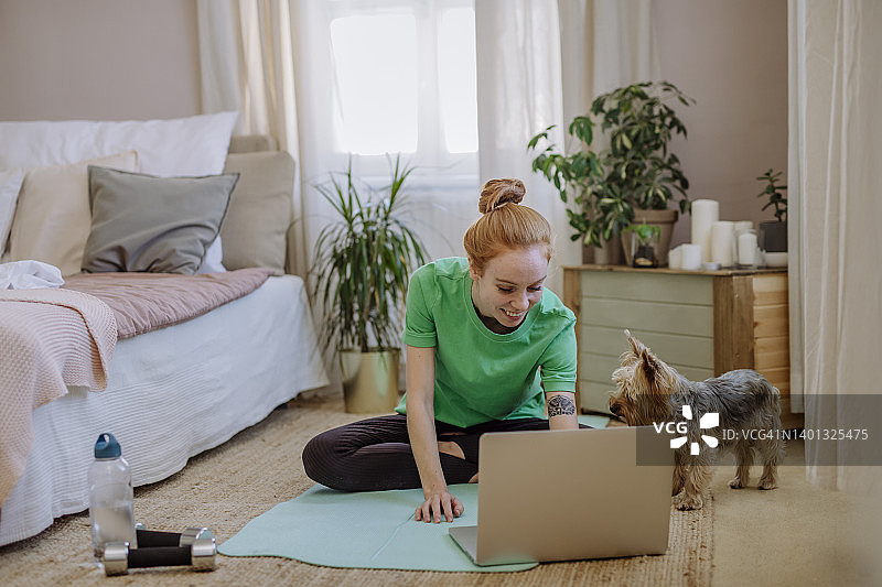 微笑的女人坐在家里宠物狗旁边的运动垫上，通过笔记本电脑观看在线教程图片素材