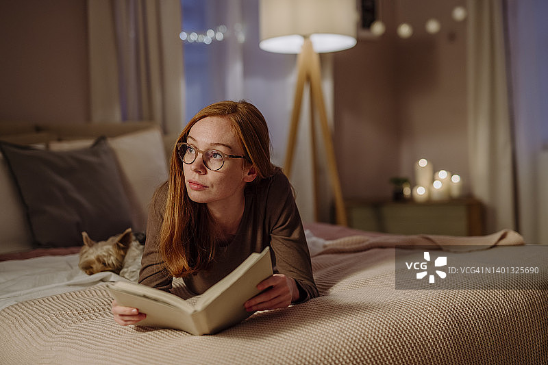 有思想的女人戴着眼镜和书躺在宠物狗在家里的床上图片素材