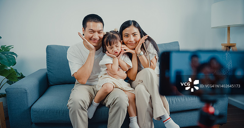 年轻的亚洲父母坐在沙发上用智能手机在家里的客厅里用摄像头打招呼。图片素材