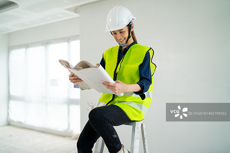 作为工程师或经理的亚裔女性在建筑工地有白墙和玻璃窗的房间里绘制房屋平面图，坐在铝梯上工作图片素材