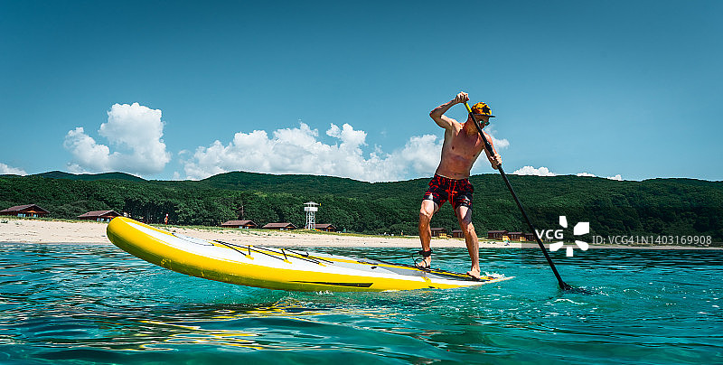晒得黝黑的男人站在黄色的冲浪板上划桨。在碧绿的海水中，沙滩上进行着积极的运动图片素材