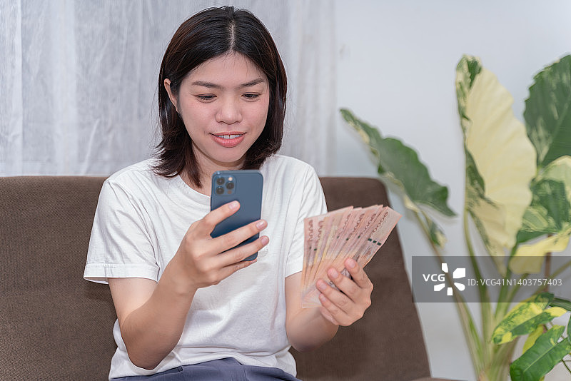 成功美丽的亚洲年轻女性手持1000泰铢纸币和智能手机，商业理念，好心情，表明他们可以很容易地借钱，现在可以实现他们的目标概念。图片素材