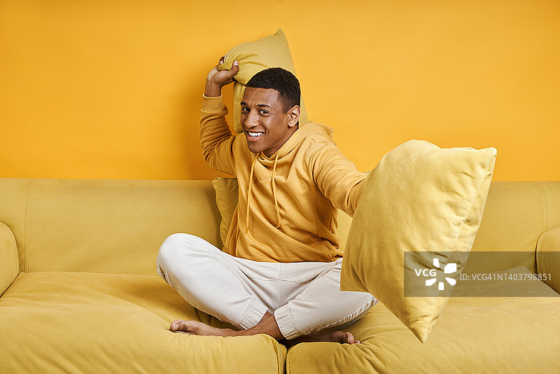 快乐的混血儿男人坐在黄色背景的沙发上准备枕头大战图片素材