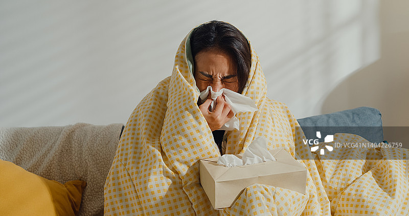 生病的年轻亚洲妇女擤鼻涕和打喷嚏用纸巾坐在毯子下的沙发在家里的客厅。图片素材