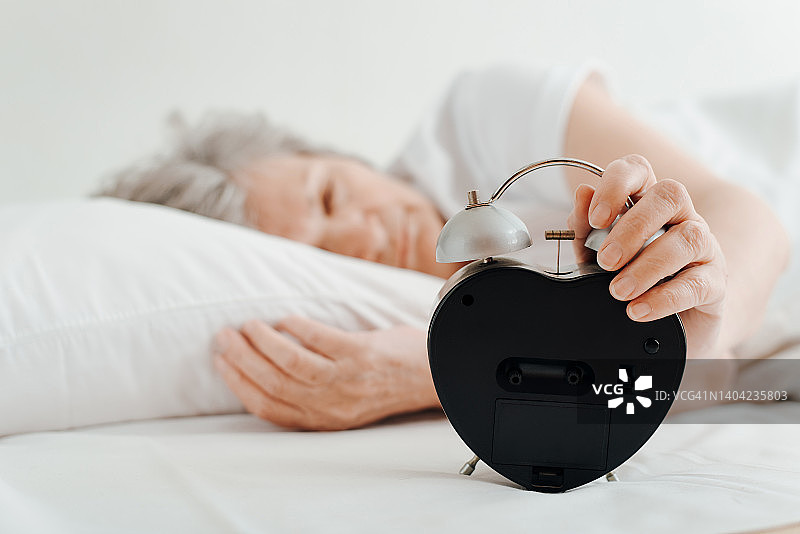 睡眼惺忪的老妇人拿着闹钟躺在床上。选择性聚焦一个闹钟和女性的手，特写。早起，清晨，老年人和睡眠观念图片素材