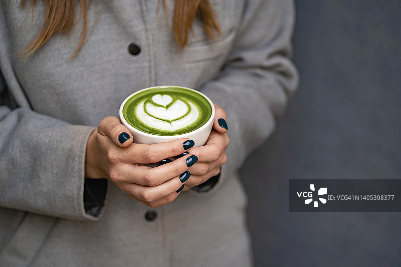 一名身穿灰色外套的女子双手捧着一个陶瓷杯，上面放着抹茶绿茶拿铁和泡沫艺术。您的设计背景与复制空间。前视图图片素材