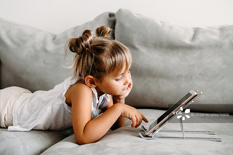 刚学步的小女孩躺在家里的沙发上玩数字无线平板电脑。宝宝的孩子成长与在线应用程序。儿童和电子设备的概念。婴儿肖像与智能手机。教育学习理念。图片素材