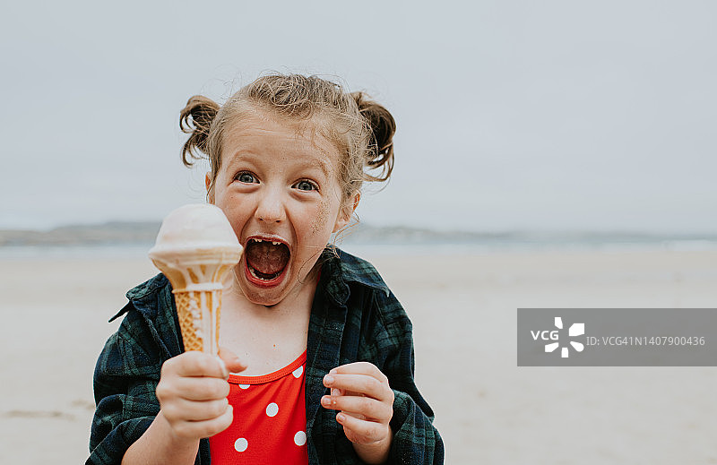 沙滩上，小女孩欣喜若狂地拿着香草冰淇淋，对着镜头拿着冰淇淋蛋筒，看起来很开心。图片素材