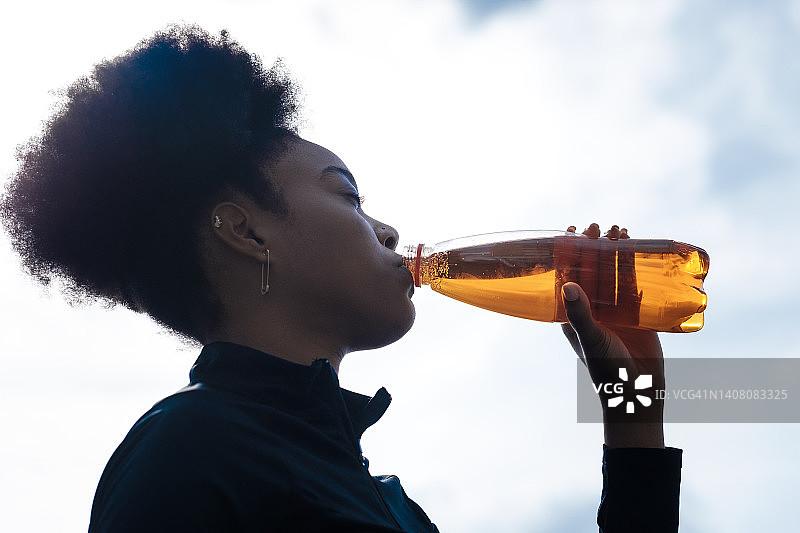 年轻健康的黑人女性的剪影喝一瓶能量饮料。图片素材