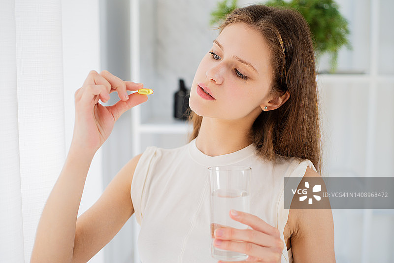 可爱的女人拿着药片Omega-3维生素和一杯水站在家里的窗边图片素材