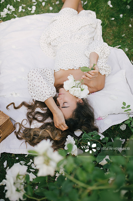 年轻美丽的女人，一头长长的卷发，穿着白色的连衣裙，在盛开的玫瑰园放松。图片素材