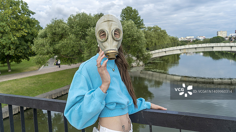 一个戴防毒面具的女孩站在河上的一座桥上。城市中的疏离感。时尚、生态、孤独的概念。图片素材