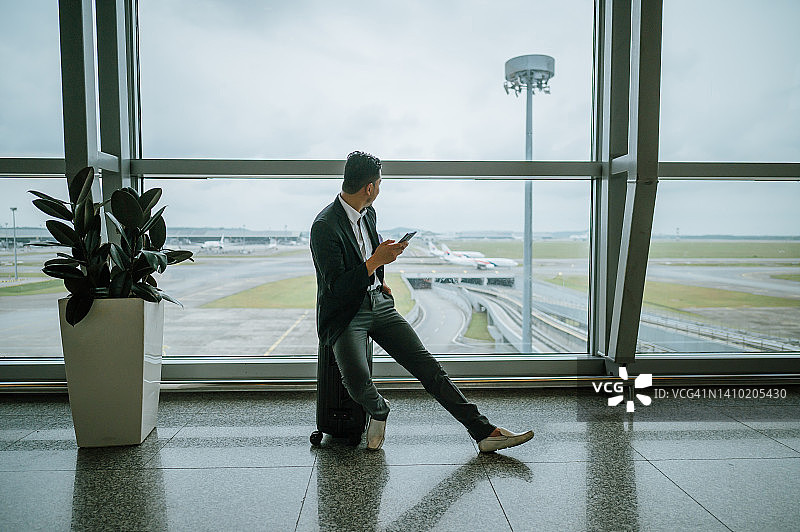 一名亚洲商人坐在行李上使用智能手机，在机场出发区图片素材