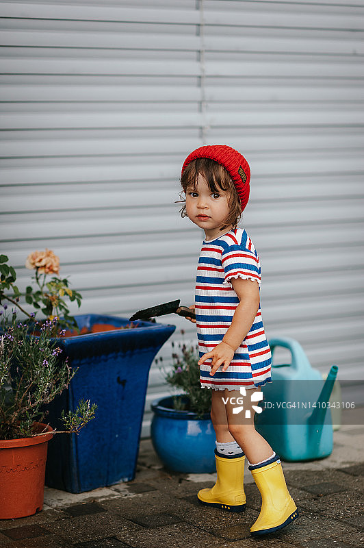 一个可爱的蹒跚学步的小女孩，戴着红色的帽子，穿着黄色的胶靴在浇花图片素材