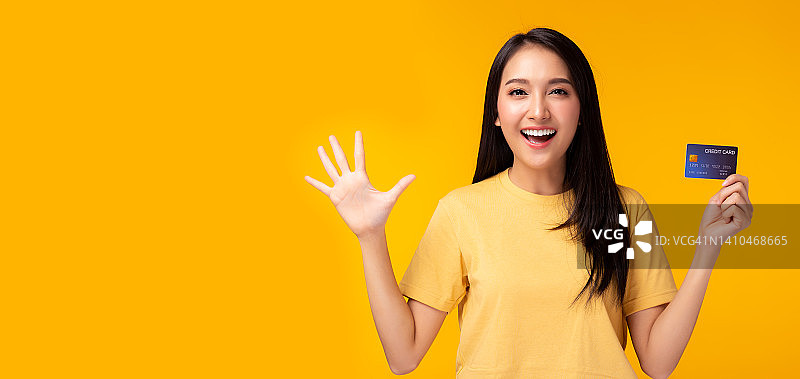 快乐的年轻亚洲女性显示信用卡和看着相机与惊讶和兴奋的脸站在黄色背景拷贝空间女孩得到满意的信用卡与幸福的微笑在脸上图片素材