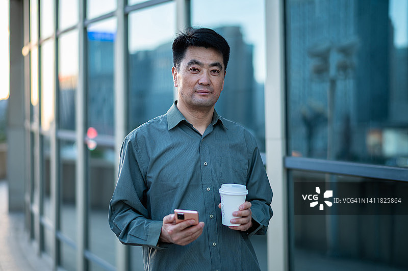 亚洲中年商人拿着咖啡杯，在城市街道上使用智能手机图片素材