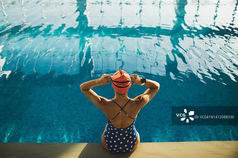 女人坐在泳池边，戴着泳镜和泳帽。从后面看。图片素材