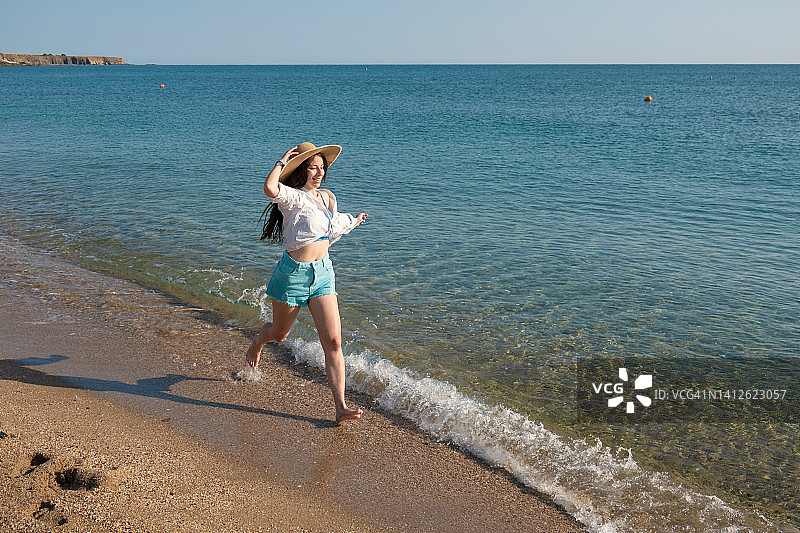 戴着草帽的年轻快乐的女人在海滩上奔跑，在浅水中嬉戏图片素材