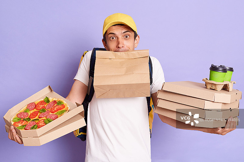 一名困惑的快递员穿着t恤，戴着帽子，手里拿着披萨盒和外卖咖啡，嘴里含着纸包站着，看起来很无助，有很多订单，在紫色背景下孤独地摆着姿势。图片素材