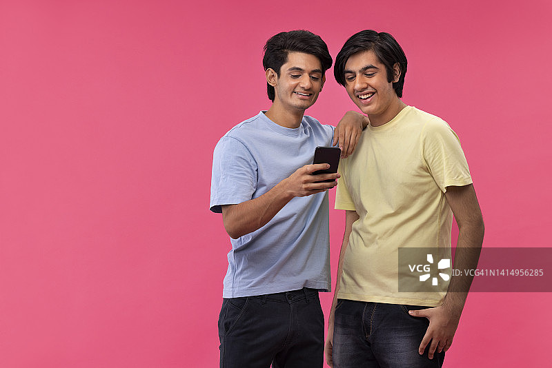 两个快乐的朋友站在粉红色的背景下用智能手机图片素材