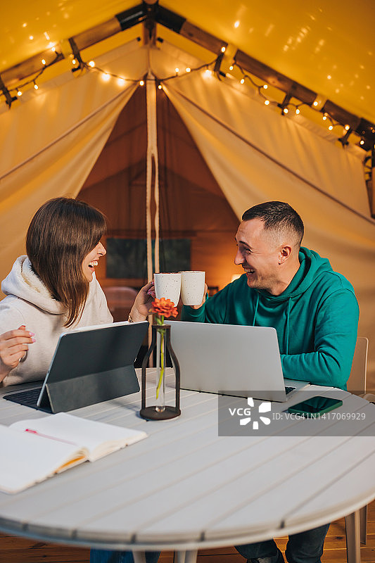 夏日傍晚，一对幸福的自由家庭夫妇在舒适的野营帐篷里用笔记本电脑工作。用于户外度假和度假的豪华露营帐篷。生活方式的概念图片素材