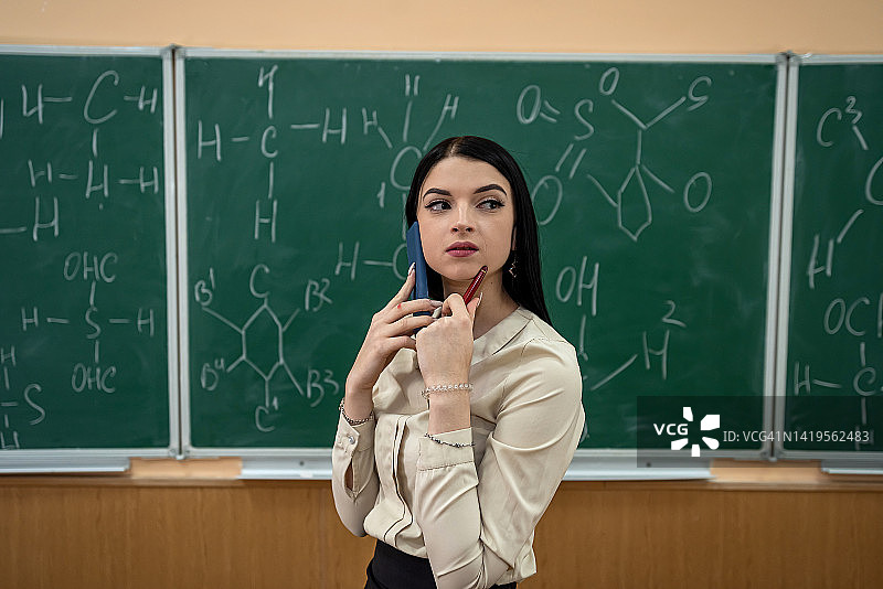 年轻漂亮的老师上化学课，站在黑板旁拿着化学式图片素材