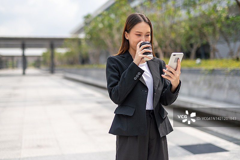 美丽年轻的亚洲女性办公室行政女士步行和使用她的智能手机和喝一杯咖啡在她的办公楼外图片素材
