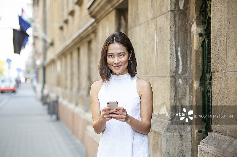 美丽的亚洲女人使用一个应用程序发送短信在她的智能手机设备。快乐的亚洲年轻女性拿着智能手机站在街上图片素材