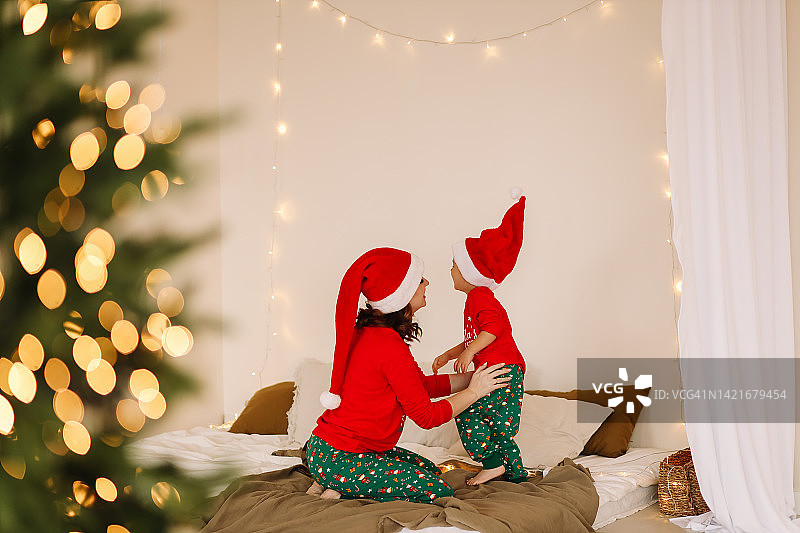 快乐的妈妈和女儿穿着红色睡衣，戴着圣诞老人的帽子，在舒适的卧室里开心地拥抱和装饰圣诞树。等待新年的到来。圣诞拍摄的概念。一个有一个孩子的家庭正在为平安夜做准备图片素材