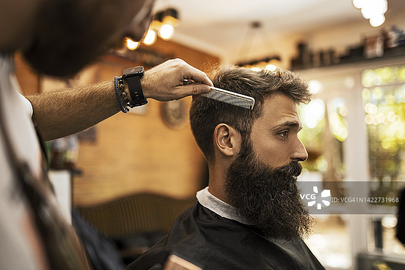 年轻的理发师正在用梳子给一个大胡子男人理发图片素材