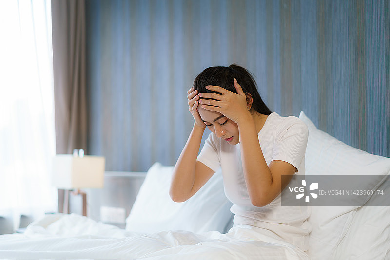 沮丧的亚洲妇女感到疲倦和头痛，坐在家里的卧室里用手捂着头。图片素材