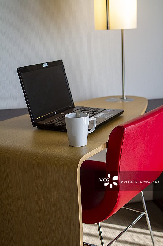 咖啡杯和笔记本电脑放在木桌上，红色的椅子聚焦在酒店房间的前景垂直照片上图片素材