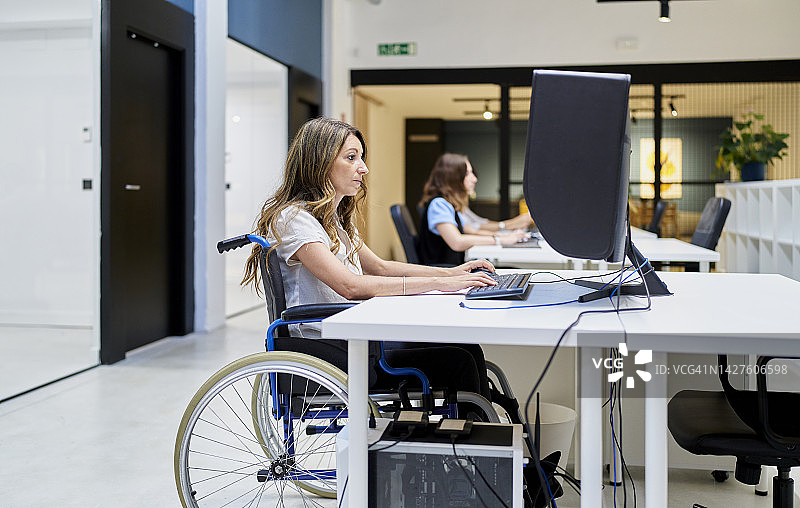 残疾妇女在办公桌前工作，坐在轮椅上，同时专注于她的日常活动在工作。图片素材