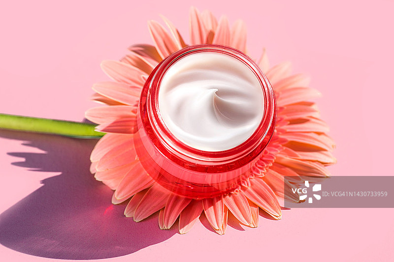 保湿霜在红粉色颜色罐和非洲菊花在柔和的粉色背景。图片素材